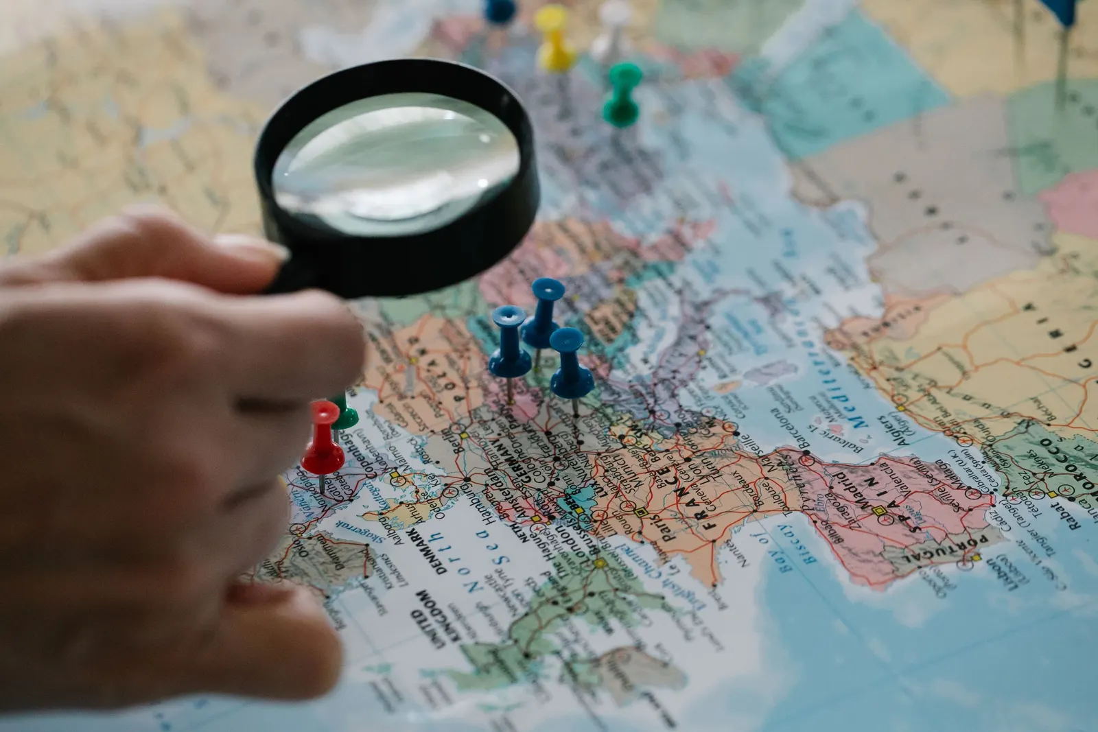 Een hand houdt een vergrootglas boven een landkaart waarop gekleurde spelden plaatsen aanduiden