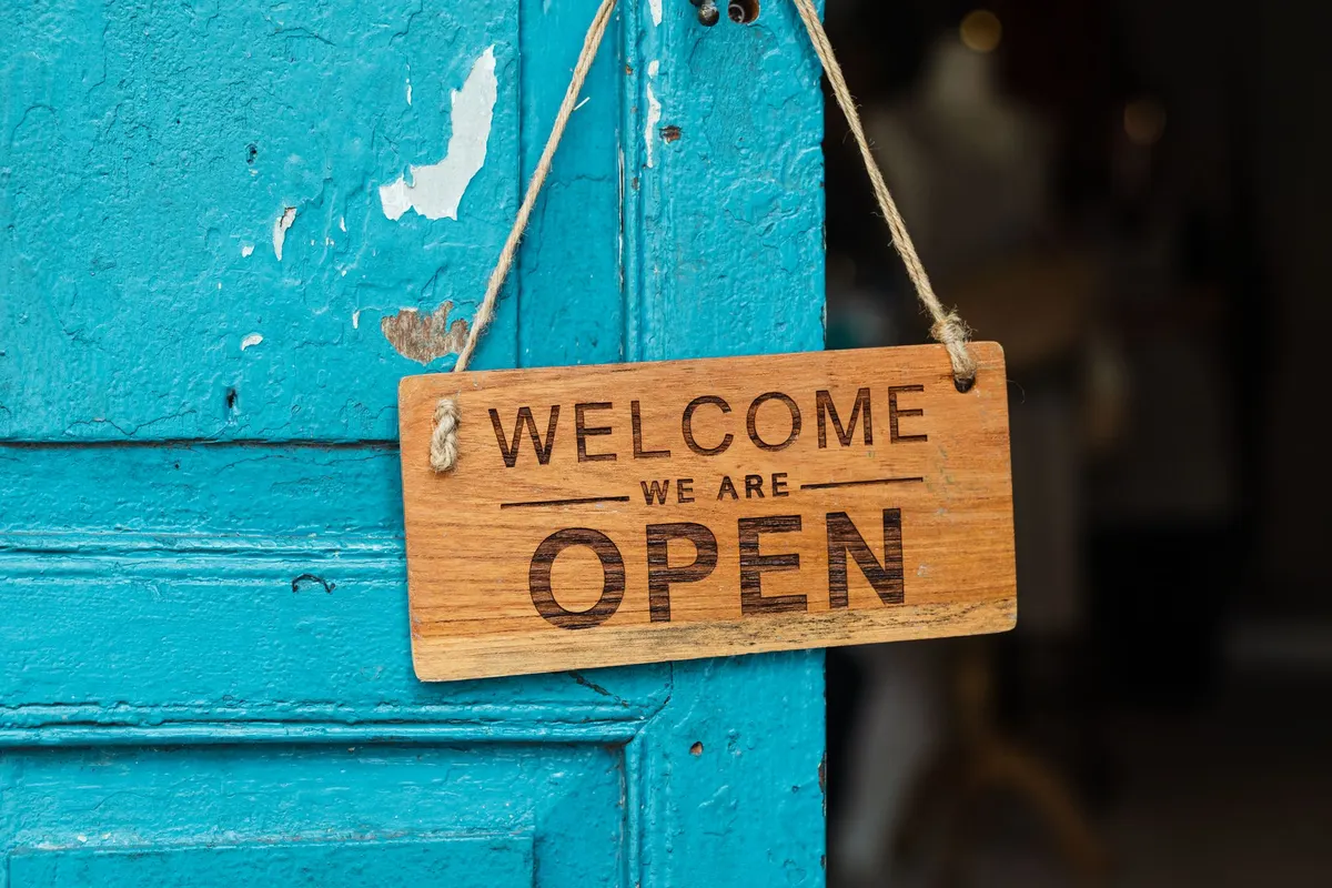 Een houten bordje met de tekst “Welcome we are open” hangt aan een turquoise deur