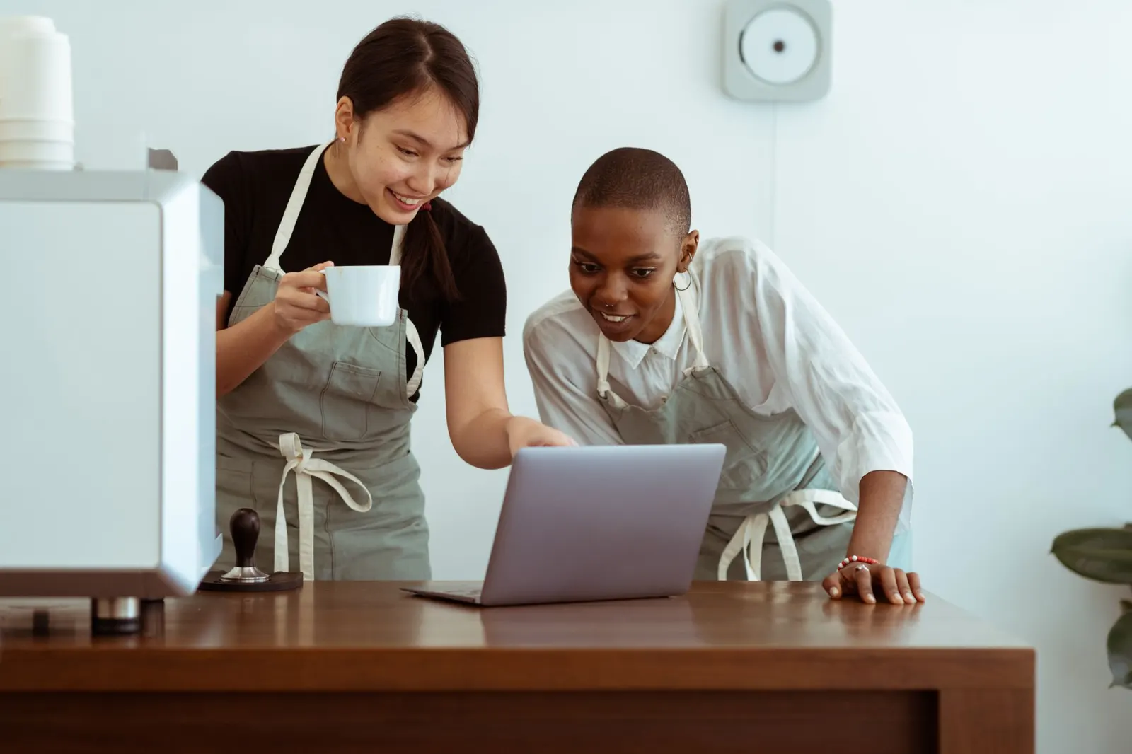 Twee vrouwen in schorten kijken lachend samen naar een laptop op de toonbank van hun zaak