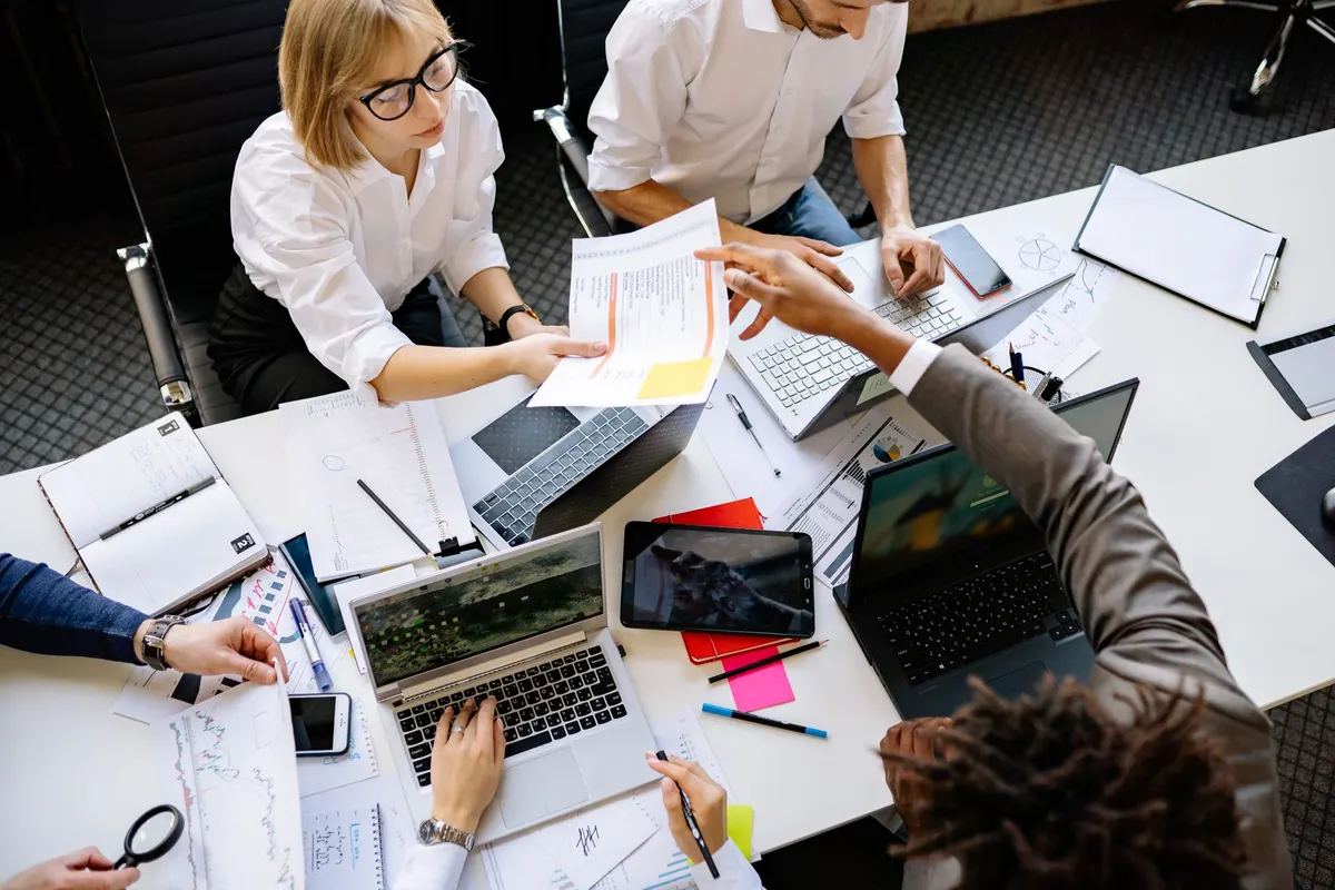 Collega’s zitten rond een tafel vol laptops, papieren en notitieboekjes, gezien van bovenaf