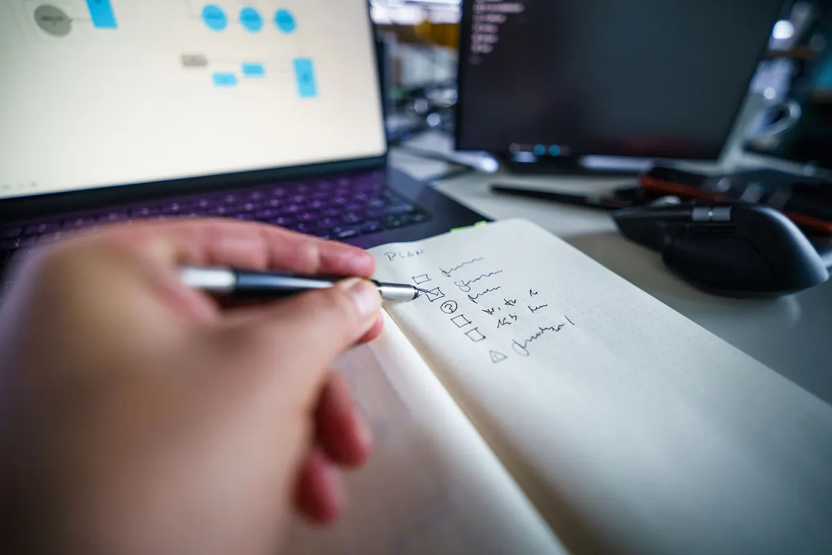 Een hand schrijft een lijstje met vakjes in een notitieboek, naast een laptop