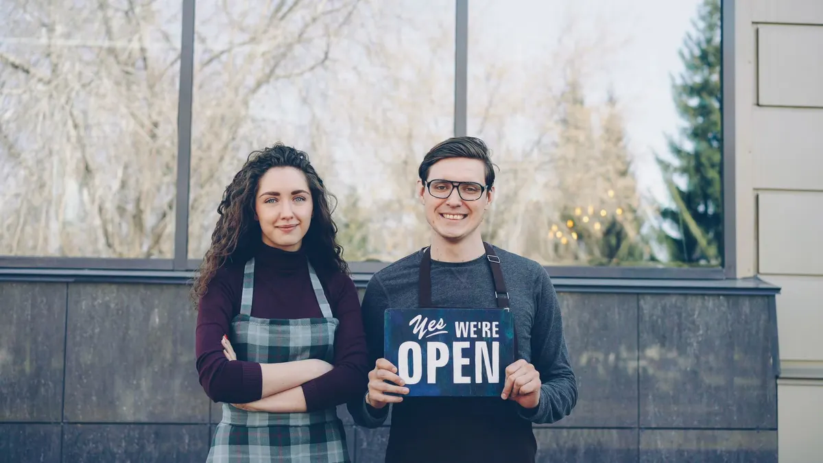 Twee ondernemers in schorten houden samen een open-bordje vast voor de ruit van hun zaak