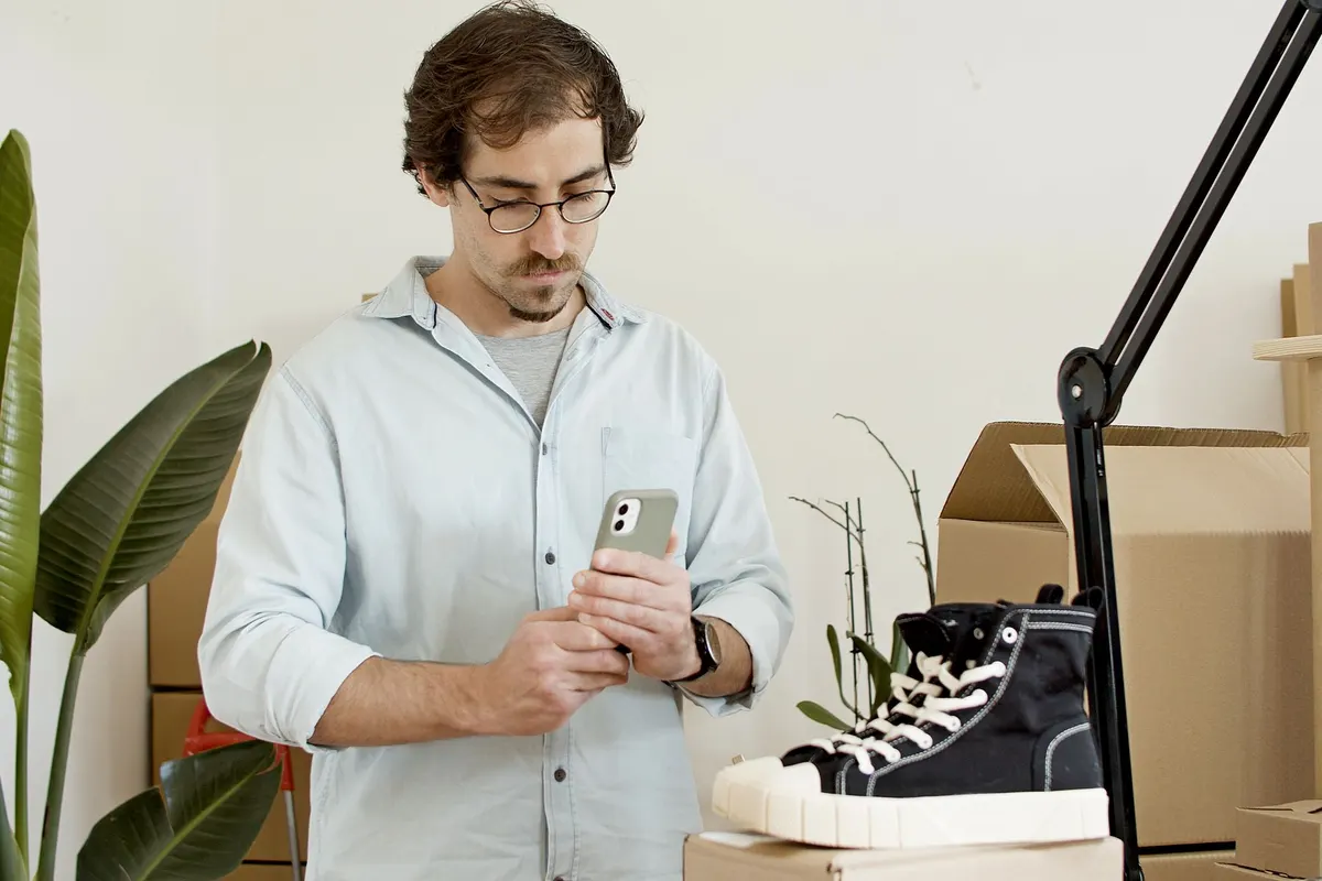 Een man in een lichtblauw hemd fotografeert met zijn smartphone een sneaker op een tafel in zijn werkruimte