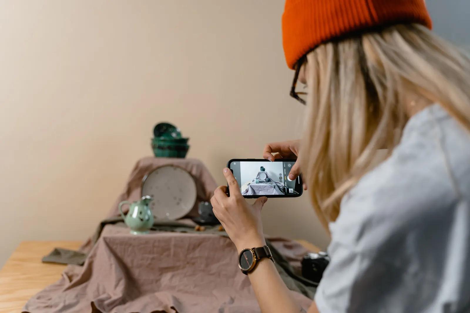 Uma mulher de gorro laranja fotografa com o celular uma louça arrumada sobre um pano