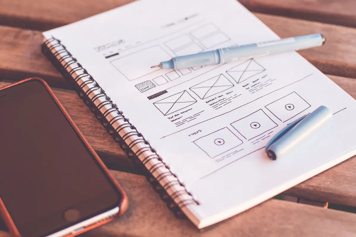 Een notitieboekje met uitgetekende schermschetsen, met een pen en een smartphone op een houten bureau