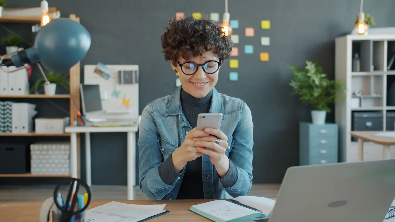 Een lachende vrouw met een bril kijkt op haar smartphone, aan een bureau met een laptop en een notitieboekje