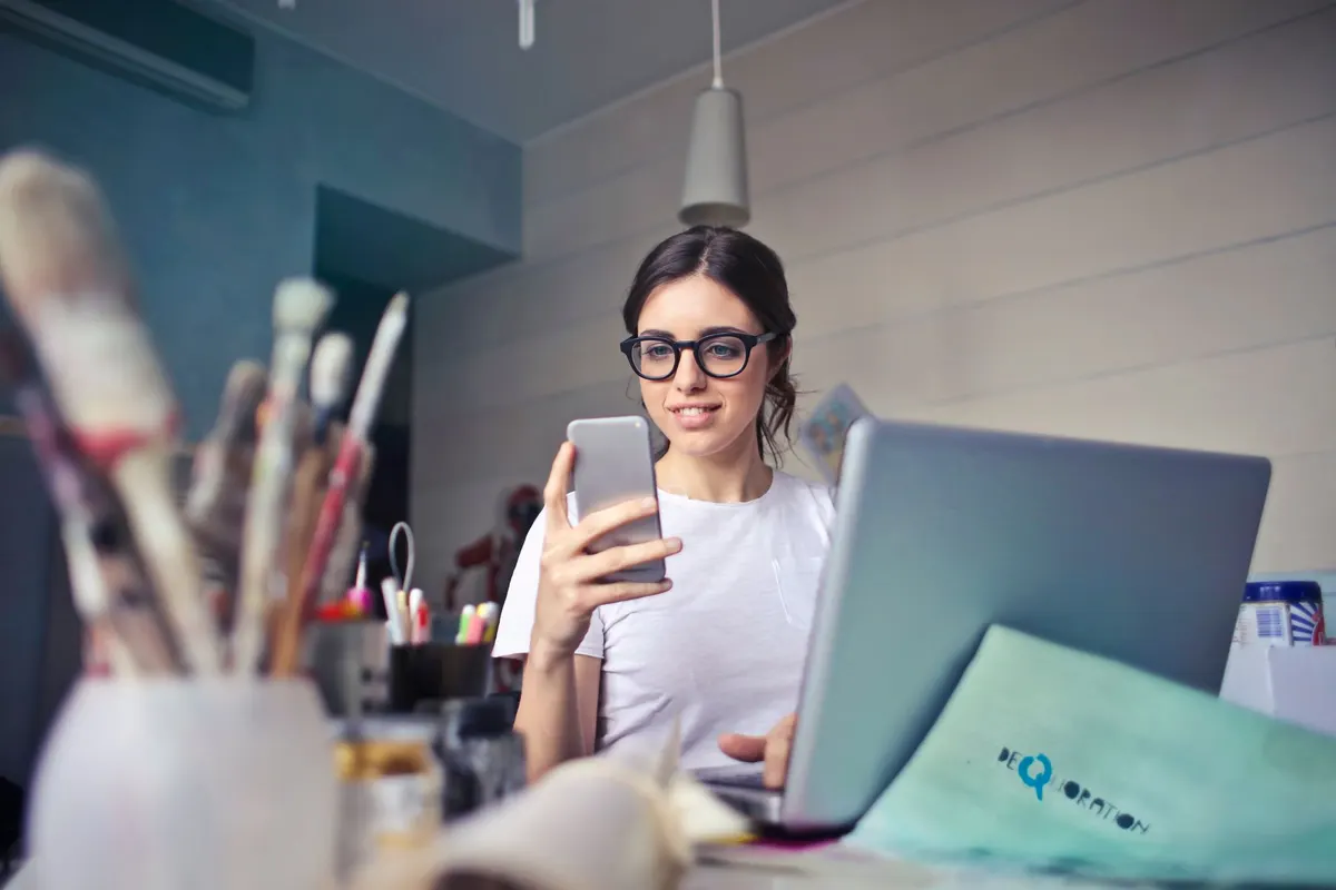 Een vrouw met een bril houdt een smartphone vast naast een laptop, tussen penselen en ander materiaal op haar bureau