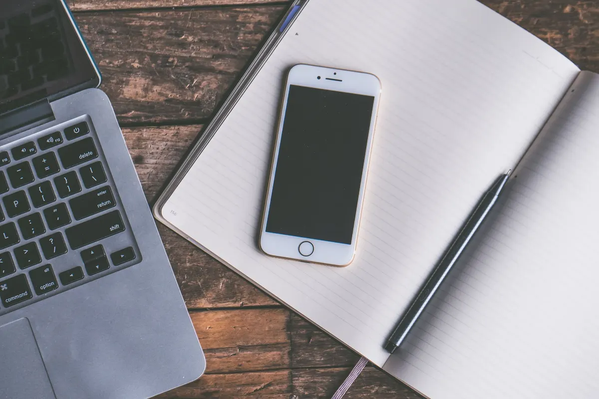 Een smartphone ligt op een open notitieboekje naast een laptop, op een houten bureau