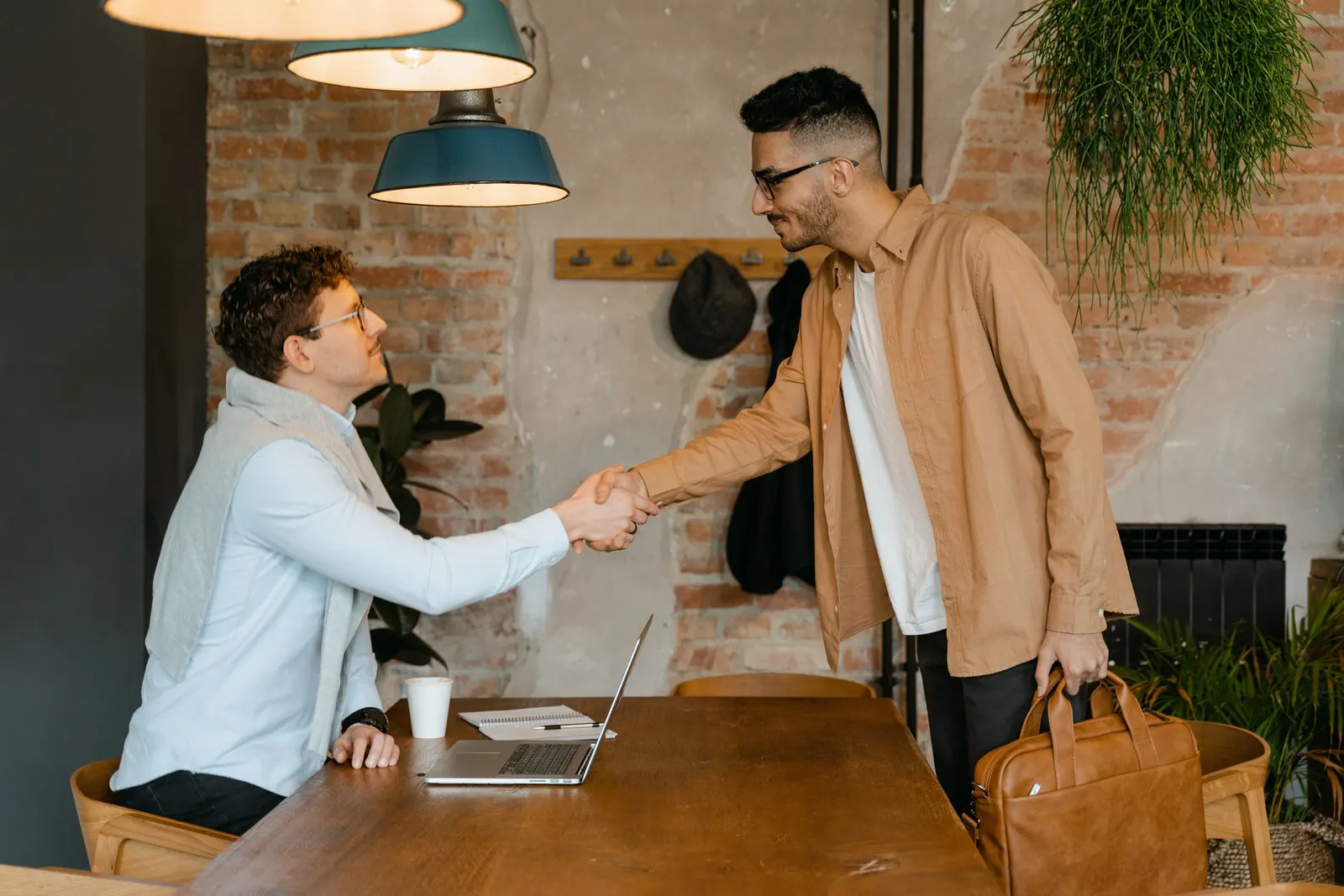 Twee mannen geven elkaar een hand aan een tafel met een laptop in een kantoor met bakstenen muur