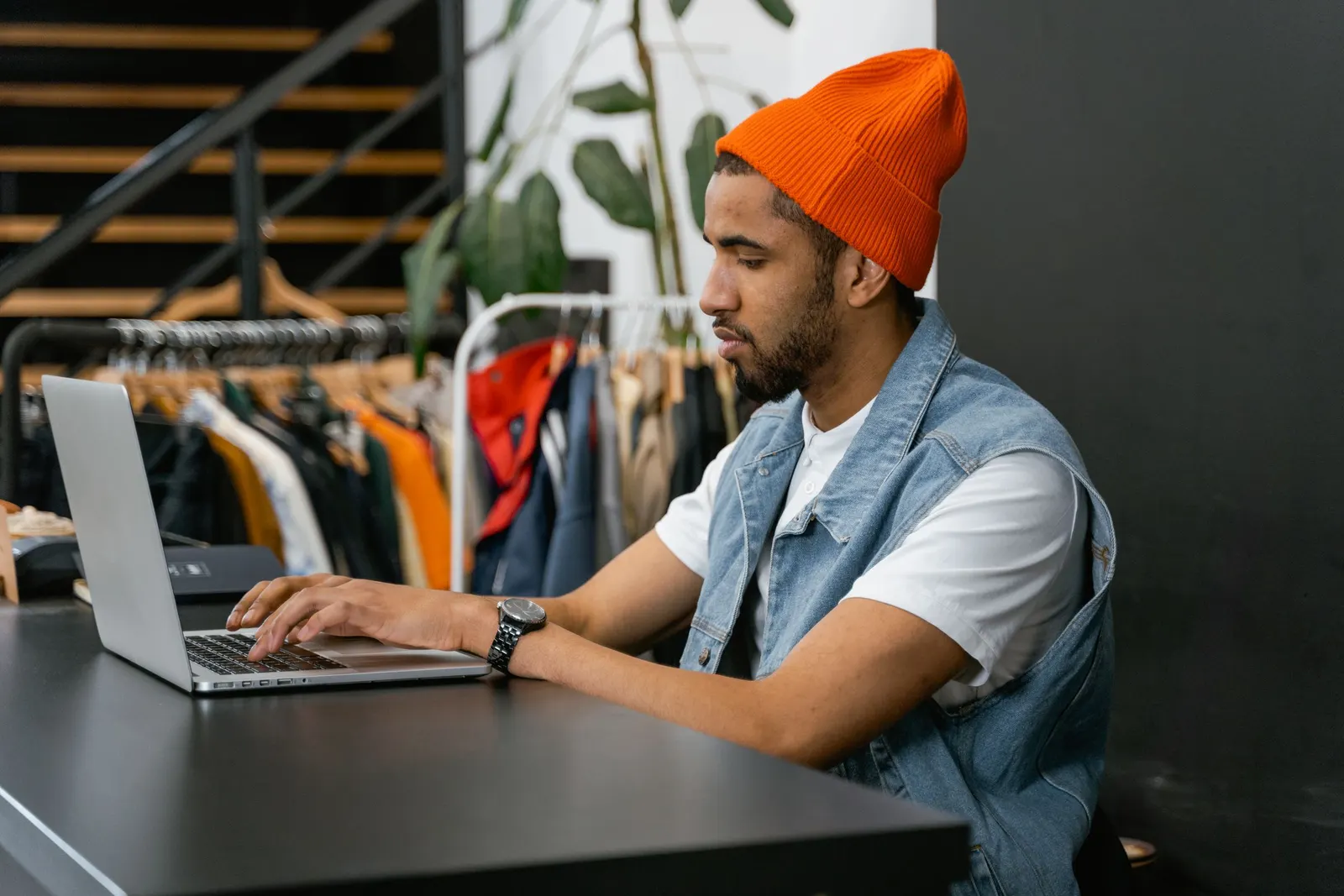 Um empreendedor de gorro laranja trabalha em um notebook no balcão da sua loja de roupas