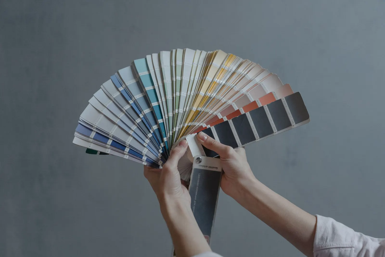 Duas mãos seguram um leque aberto de amostras de cores diante de um fundo cinza
