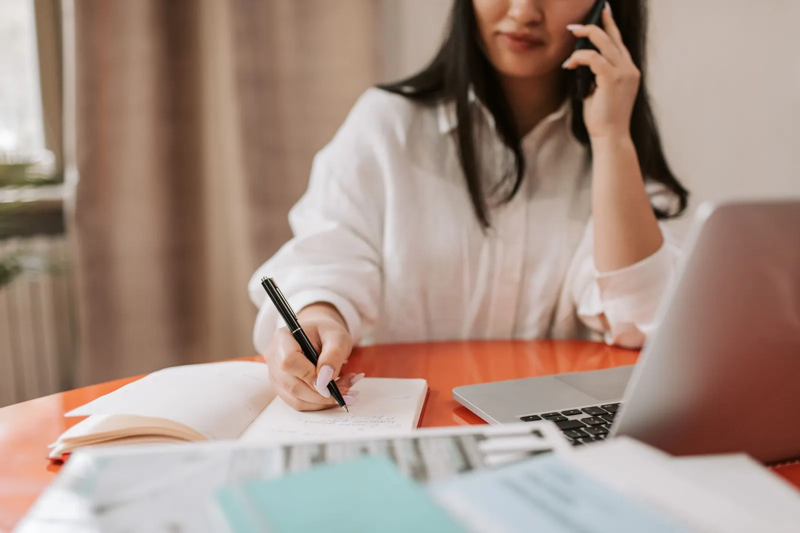 Een vrouw belt aan een bureau met een laptop en schrijft tegelijk notities in een notitieboek