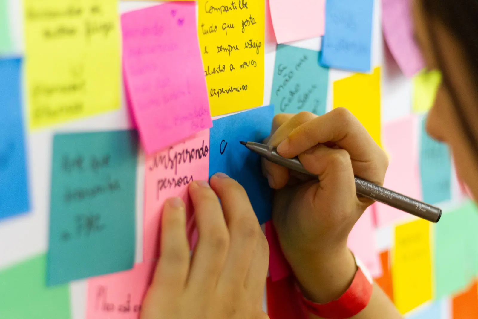 Uma mão escreve com uma caneta em um post-it, no meio de muitos post-its coloridos com palavras em uma parede