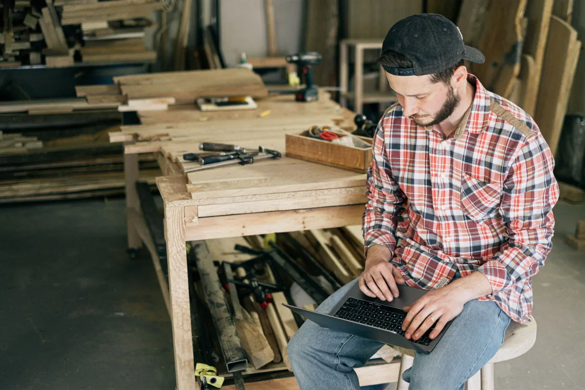 Een vakman werkt op zijn laptop in zijn houtwerkplaats, tussen het gereedschap