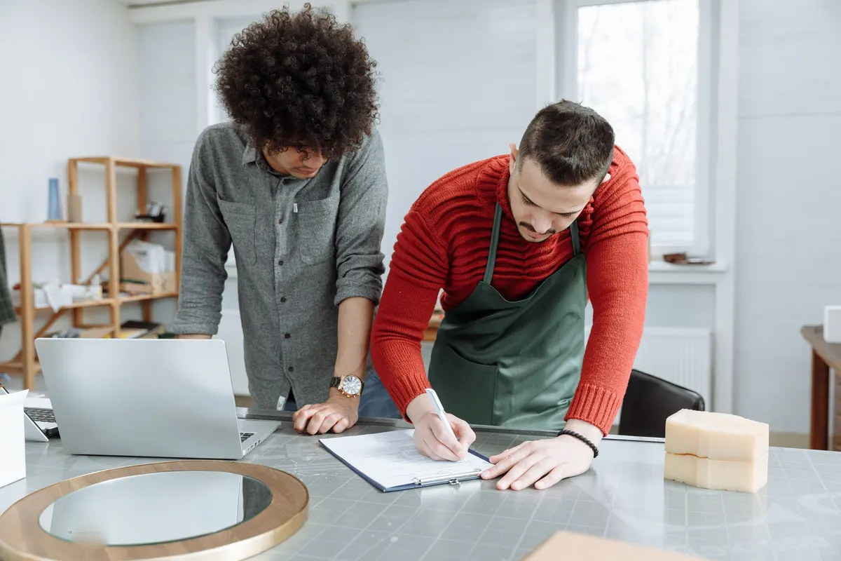 Een ondernemer in werkschort neemt met iemand aan tafel een plan door, met een laptop en notities