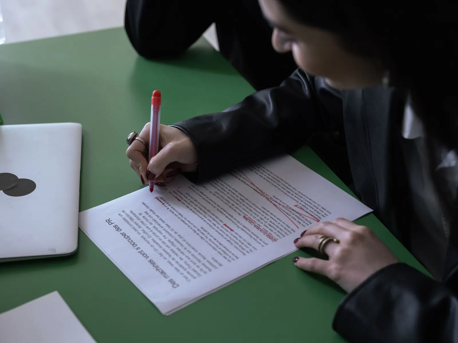 Iemand verbetert een afgedrukte tekst met een rode pen op een groene tafel, naast een laptop