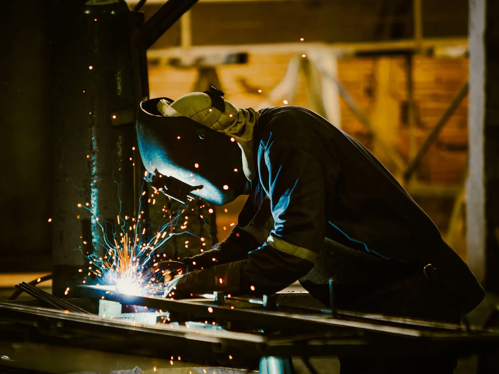 Een lasser met beschermkap en handschoenen last een metalen stuk in een werkplaats, met rondvliegende vonken