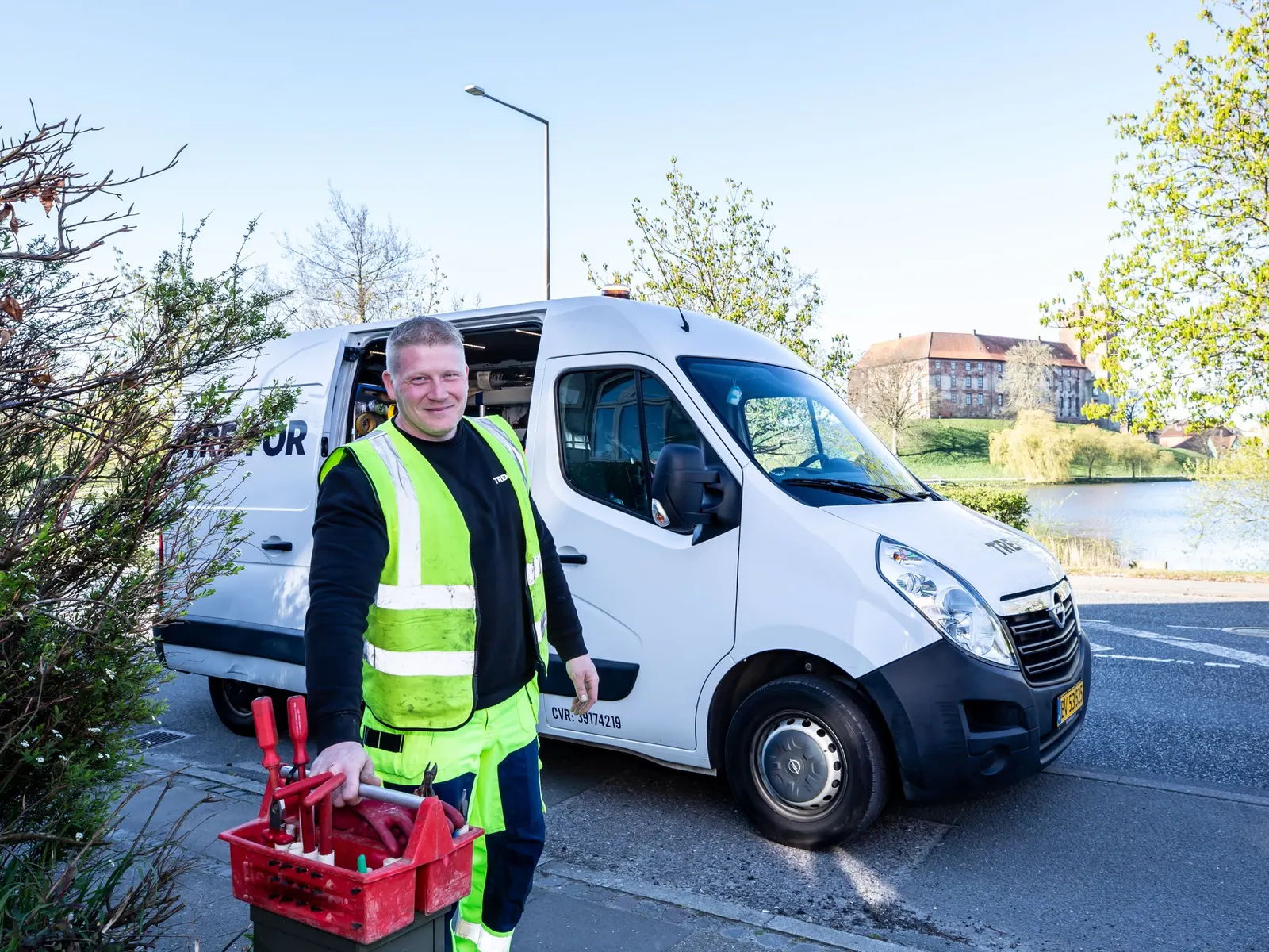 Een lachende vakman in een fluohesje staat met zijn gereedschap naast een witte bestelwagen