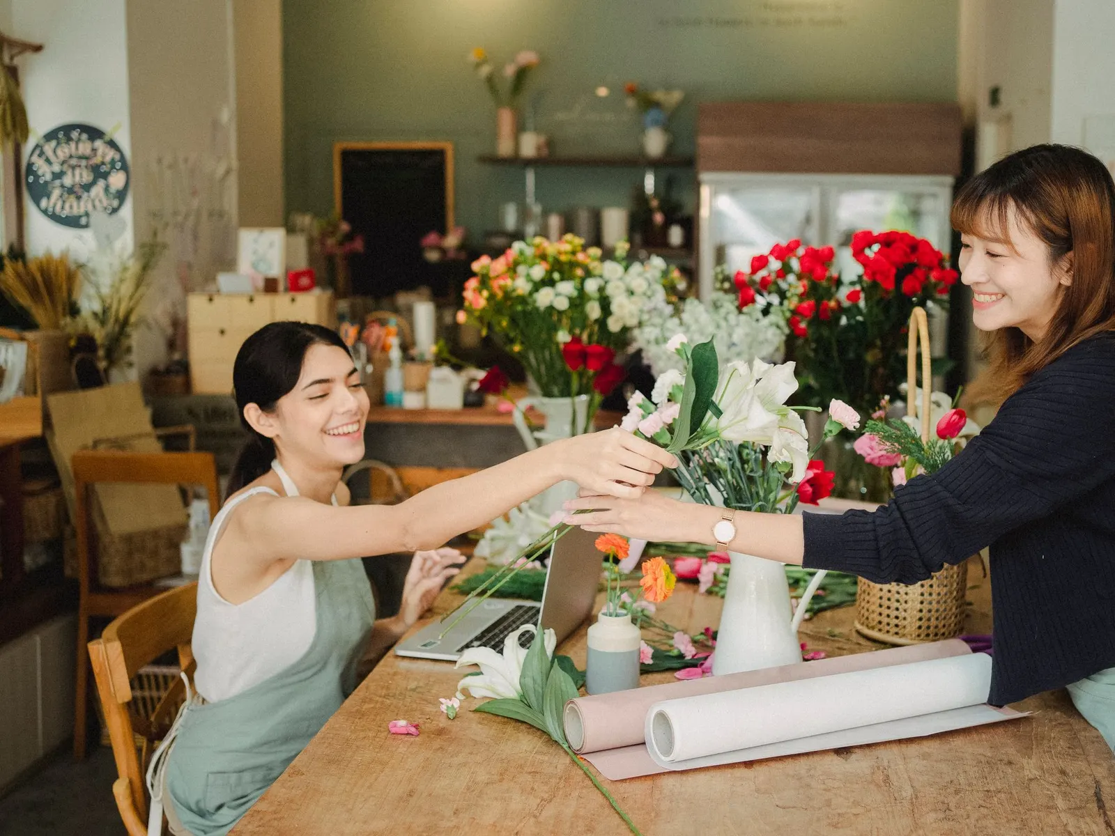 Twee vrouwen in een bloemenwinkel geven elkaar een boeket door, met een laptop op de werktafel